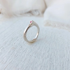 May include: A silver ring with a small, round, pink gemstone. The ring has a textured, organic design. The gemstone is set at the top of the ring. The ring is displayed on a white surface.
