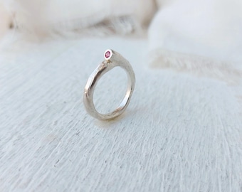 Puede incluir: Un anillo de plata con una pequeña gema rosa y redonda. El anillo tiene un diseño texturizado y orgánico. La gema está engastada en la parte superior del anillo. El anillo se muestra sobre una superficie blanca.
