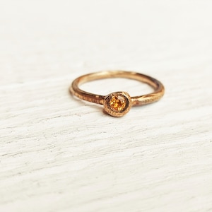 Op de afbeelding: Een goudkleurige ring met een ronde, oranje edelsteen gezet in een getextureerde bezel. De band heeft een gehamerde uitstraling. De ring wordt gepresenteerd op een wit, getextureerd oppervlak. Een sieraad.