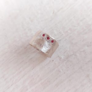 Puede incluir: Un anillo plateado texturizado con tres pequeñas piedras rojas redondas incrustadas. El anillo tiene una banda ancha y una superficie ligeramente irregular, lo que le da un aspecto artesanal. Las piedras están espaciadas uniformemente.