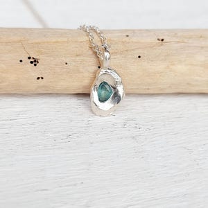 Peut inclure: Un collier pendentif en argent avec un pendentif de forme organique et rugueuse. Le pendentif contient une petite pierre précieuse de couleur teal. La chaîne est également en argent. Le bijou est présenté sur un morceau de bois flotté sur un fond en bois blanc.