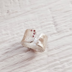 Op de afbeelding: Een zilveren ring met een getextureerd, open ontwerp en kleine rode edelstenen.