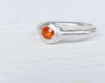 Könnte beinhalten: Ein silberner Ring mit einem runden, orangefarbenen Edelstein, der in einer strukturierten Lünette gefasst ist. Der Ring hat ein schlichtes Band und ein leicht gehämmertes Aussehen, das eine handwerkliche Ästhetik zeigt. Der Edelstein hat eine warme Farbe.