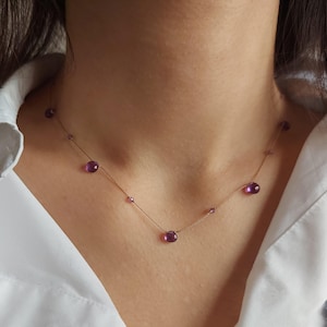 Puede incluir: Un delicado collar con varias piedras preciosas redondas de color púrpura intenso ensartadas en una fina cadena dorada. El collar se lleva sobre una camisa blanca.