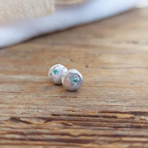 Puede incluir: Un par de pendientes de plata con un acabado texturizado y una pequeña piedra turquesa en el centro de cada pendiente.