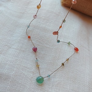 May include: A delicate necklace featuring a variety of colorful gemstone beads strung on a thin, brown cord. The beads include shades of pink, yellow, orange, green, and blue. The necklace is displayed on a white, textured surface.