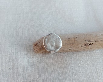 May include: A silver ring with a round, textured top. The ring is sitting on a piece of driftwood.