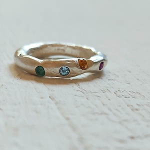 Peut inclure: Une bague en argent à la surface texturée, ornée d'une série de pierres précieuses colorées. La bague a une forme irrégulière unique et est ornée de pierres vertes, bleues, orange et roses. Un bijou.