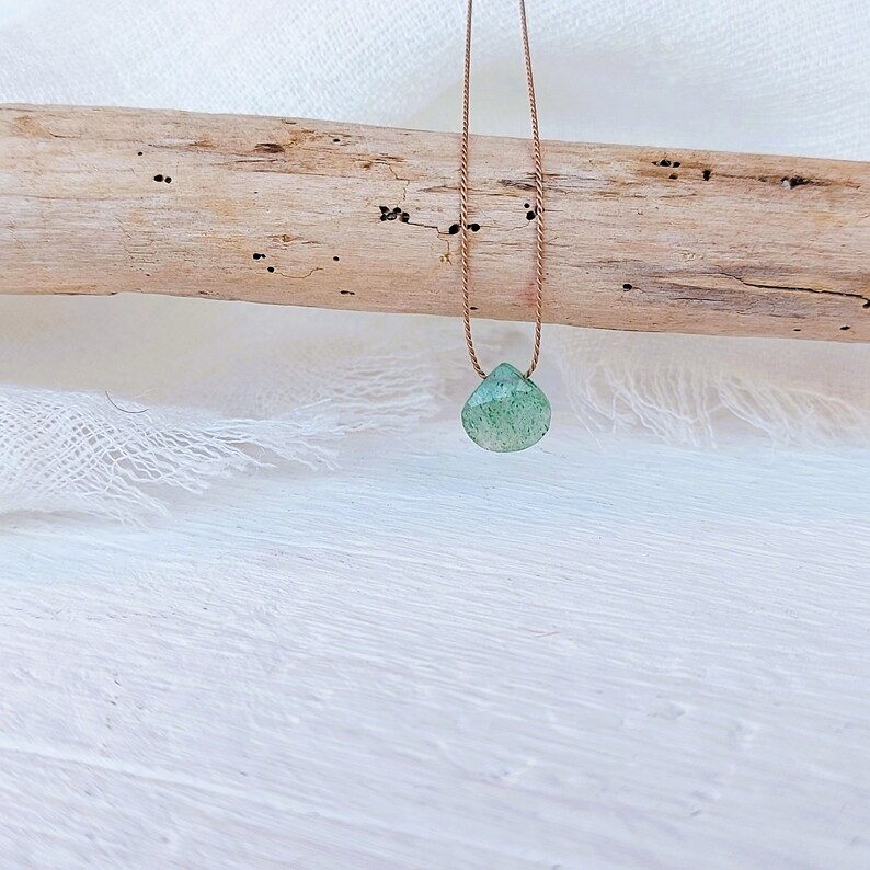 May include: A green gemstone pendant on a brown cord necklace. The pendant is a teardrop shape and has a speckled appearance.