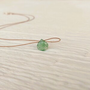 May include: A delicate green gemstone pendant on a brown cord necklace. The pendant is a teardrop shape and has a natural, rough texture.