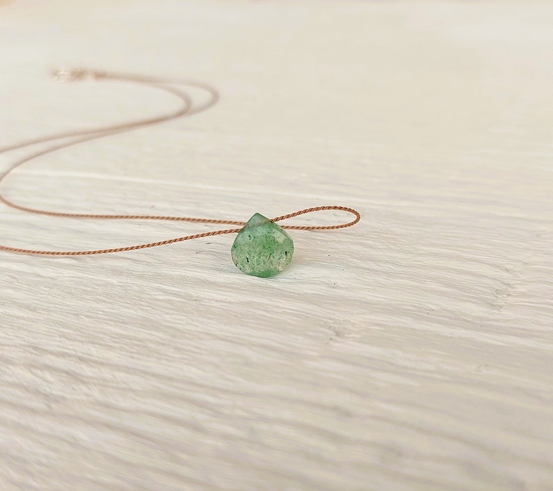 May include: A delicate green gemstone pendant on a brown cord necklace. The pendant is a teardrop shape and has a natural, rough texture.