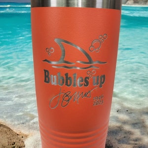 May include: Orange stainless steel tumbler with a shark fin and bubbles design. The text "Bubbles Up" is engraved on the tumbler, along with a signature and the dates 1946 and 2023.