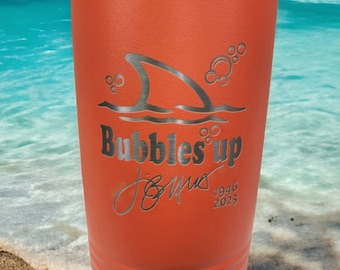May include: Orange stainless steel tumbler with a shark fin and bubbles design. The text "Bubbles Up" is engraved on the tumbler, along with a signature and the dates 1946 and 2023.