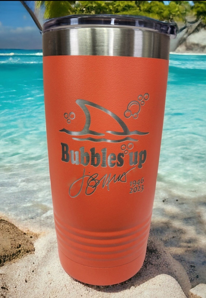 May include: Orange stainless steel tumbler with a shark fin and bubbles design. The text "Bubbles Up" is engraved on the tumbler, along with a signature and the dates 1946 and 2023.