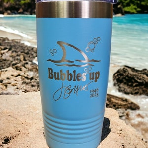 May include: A light blue stainless steel tumbler with a shark fin and bubbles design. The tumbler has the text "Bubbles Up" and a signature with the dates 1946 and 2023.