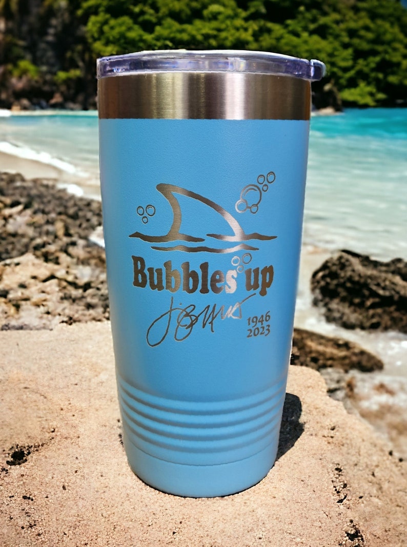 May include: A light blue stainless steel tumbler with a shark fin and bubbles design. The tumbler has the text "Bubbles Up" and a signature with the dates 1946 and 2023.