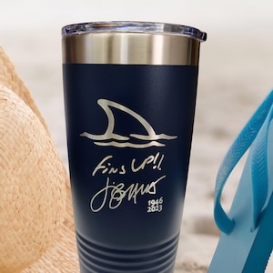 May include: Navy blue insulated tumbler with a silver rim and lid. The tumbler features a shark fin graphic and the text "Fins Up!!" along with a signature and the years 1946-2023. A beach theme is suggested by the background.