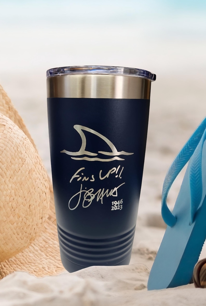 May include: Navy blue insulated tumbler with a silver rim and lid. The tumbler features a shark fin graphic and the text "Fins Up!!" along with a signature and the years 1946-2023. A beach theme is suggested by the background.