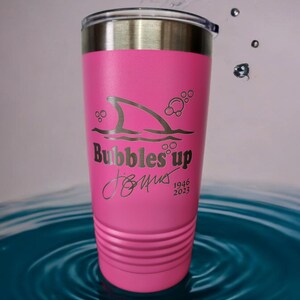 May include: Pink stainless steel tumbler with a shark fin and bubbles design. The tumbler has the text "Bubbles Up" and a signature with the dates 1946 and 2023.
