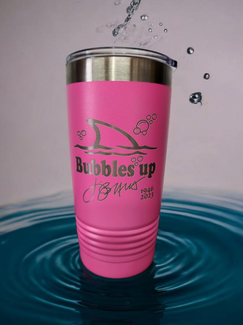 May include: Pink stainless steel tumbler with a shark fin and bubbles design. The tumbler has the text "Bubbles Up" and a signature with the dates 1946 and 2023.