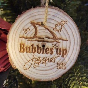 May include: A wooden Christmas ornament with a shark fin and bubbles, the text "Bubbles'up" and the year "1946 2023".