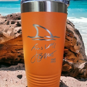 May include: Orange insulated tumbler with a silver shark fin design and the words "Fins Up!! Jones 1946 2023". The tumbler has a silver rim and a clear lid. The tumbler is sitting on a sandy surface.