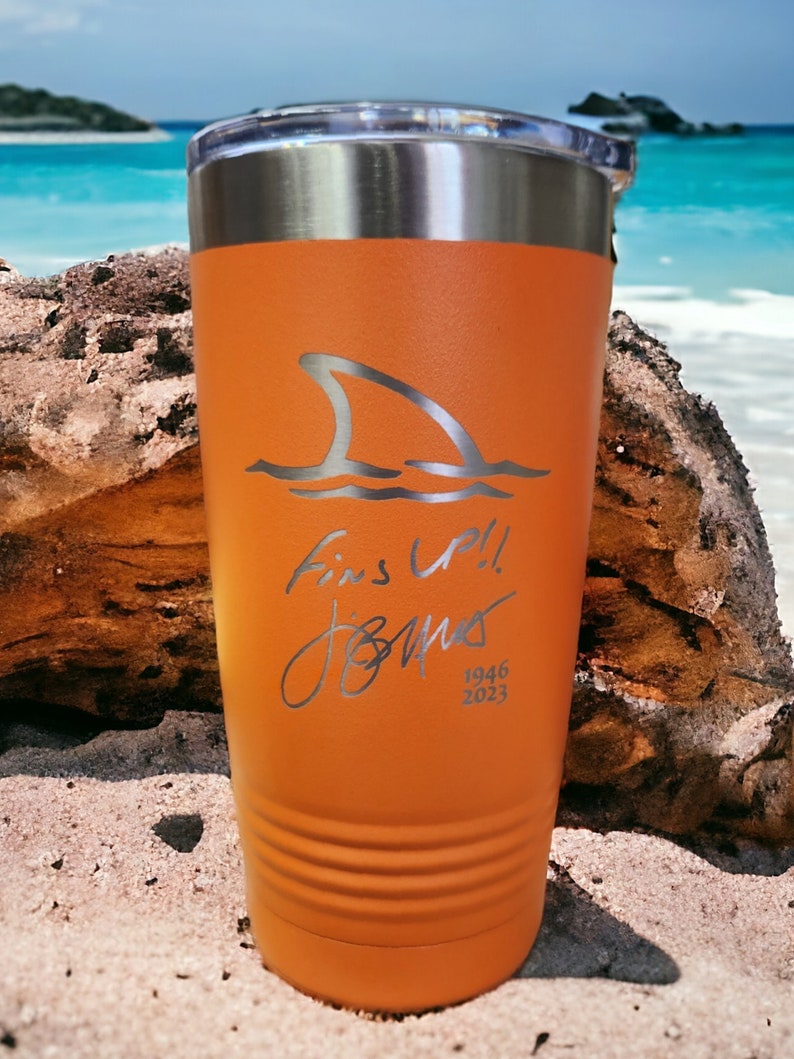 May include: Orange insulated tumbler with a silver shark fin design and the words "Fins Up!! Jones 1946 2023". The tumbler has a silver rim and a clear lid. The tumbler is sitting on a sandy surface.