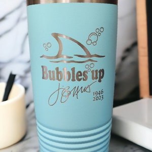 May include: A light blue stainless steel tumbler with a shark fin and bubbles design. The tumbler has a silver lid and says "Bubbles Up" and "Jimmy 1946 2023".