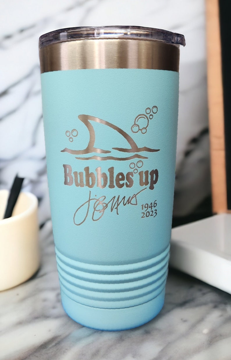May include: A light blue stainless steel tumbler with a shark fin and bubbles design. The tumbler has a silver lid and says "Bubbles Up" and "Jimmy 1946 2023".