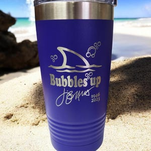 May include: A purple stainless steel tumbler with a shark fin and bubbles design. The tumbler has the text "Bubbles Up" and "Jimus 1946 2023" engraved on it.