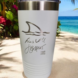 May include: White insulated tumbler with a silver lid. The tumbler features a shark fin graphic with the words "Fins Up!!" and a signature, along with the numbers "1946 2023". The background shows a beach scene.