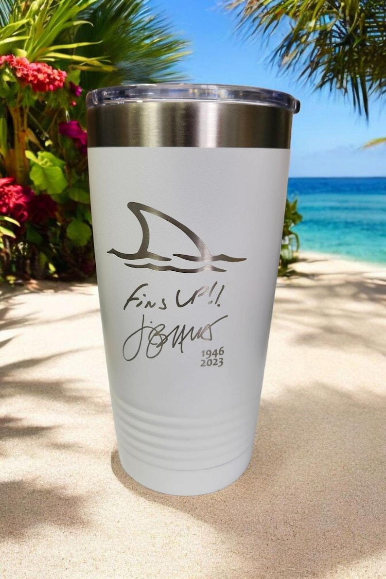 May include: White insulated tumbler with a silver lid. The tumbler features a shark fin graphic with the words "Fins Up!!" and a signature, along with the numbers "1946 2023". The background shows a beach scene.