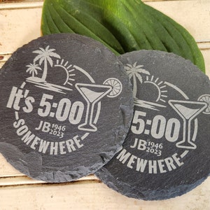 May include: Two black slate coasters with a white design. The design includes a palm tree, a sun, a cocktail glass, and the text "It's 5:00 Somewhere JB 1946-2023".