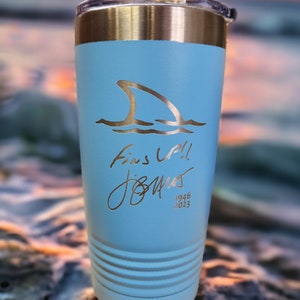May include: A light blue insulated tumbler with a gold-colored shark fin design and the words "Fins Up!!" and a signature. The tumbler has a silver rim and a black lid. The year 1946-2023 is printed below the signature.