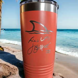 May include: Coral-colored travel tumbler with a shark fin graphic and the text "Fins Up! Jo Muir 1946-2023". The stainless steel tumbler has a black lid and is suitable for hot or cold beverages.