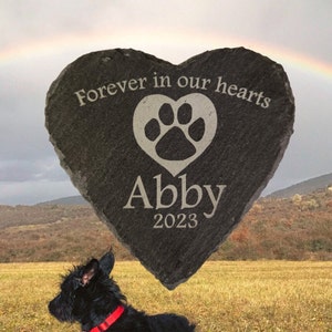 May include: A black heart-shaped stone memorial with the text "Forever in our hearts" and a paw print inside a heart. The name "Abby" and the year "2023" are engraved below the heart. A black dog is in the foreground.