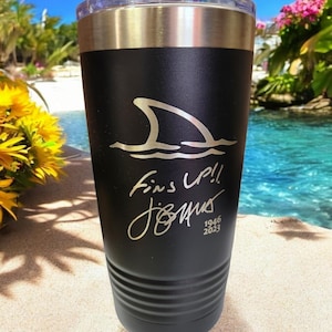May include: Black insulated tumbler with a silver rim and clear lid. The tumbler features a shark fin design above the words "Fins Up!!" and a signature. The bottom of the tumbler has a ridged design. The text "1946 2023" is also visible.