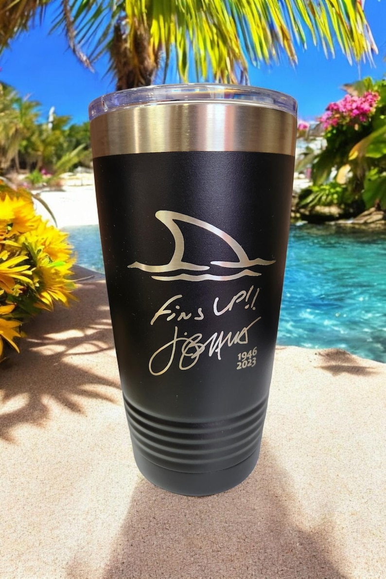 May include: Black insulated tumbler with a silver rim and clear lid. The tumbler features a shark fin design above the words "Fins Up!!" and a signature. The bottom of the tumbler has a ridged design. The text "1946 2023" is also visible.