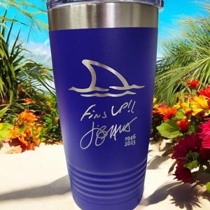 May include: A blue insulated tumbler with a silver rim and lid. The tumbler features a shark fin graphic above the words "Fins Up!!" and a signature, with the years "1946 2023" below. The background shows a beach scene.