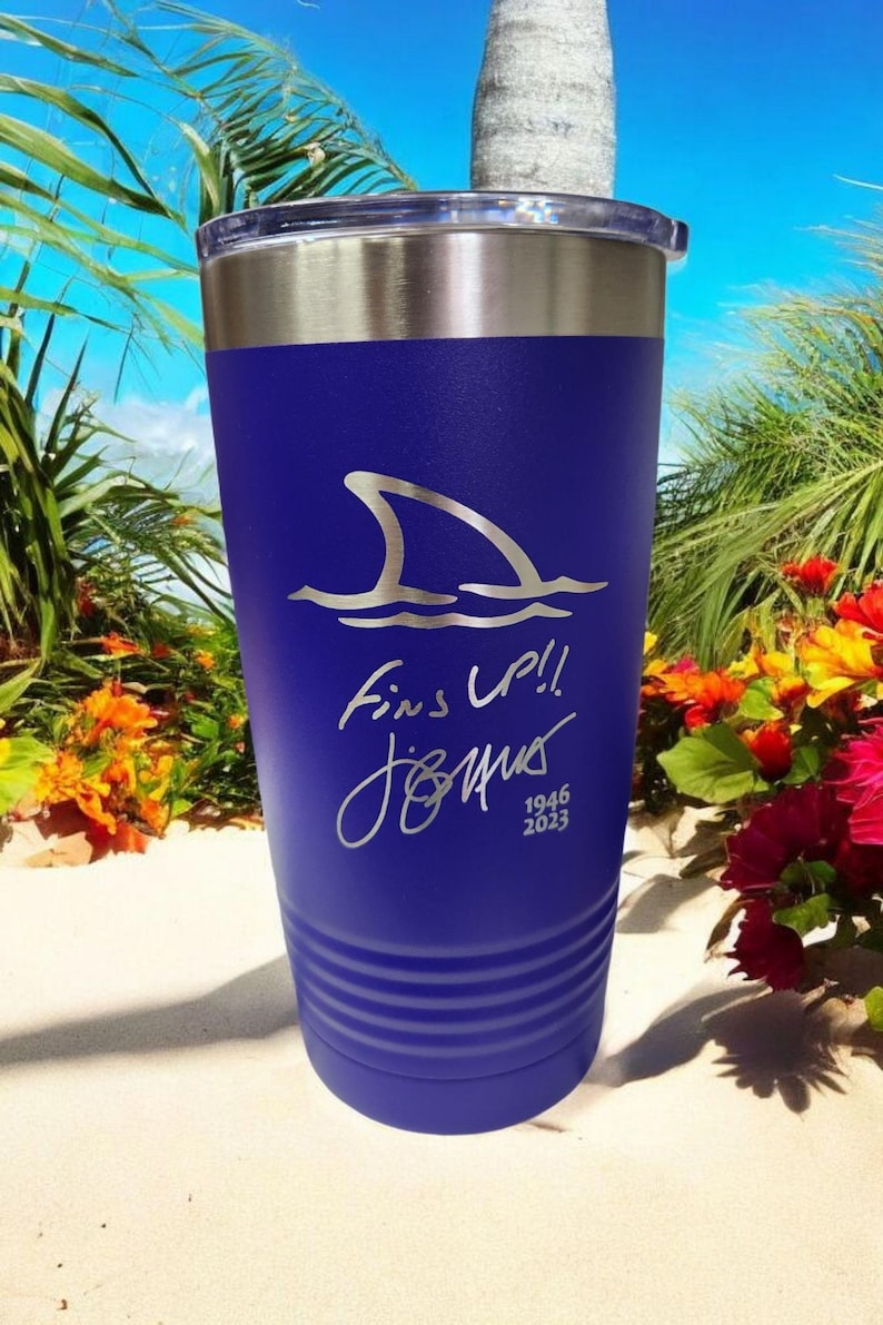 May include: A blue insulated tumbler with a silver rim and lid. The tumbler features a shark fin graphic above the words "Fins Up!!" and a signature, with the years "1946 2023" below. The background shows a beach scene.