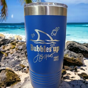 May include: A blue stainless steel tumbler with a shark fin and bubbles design. The tumbler is engraved with the text "Bubbles Up" and a signature with the dates 1946 and 2023.