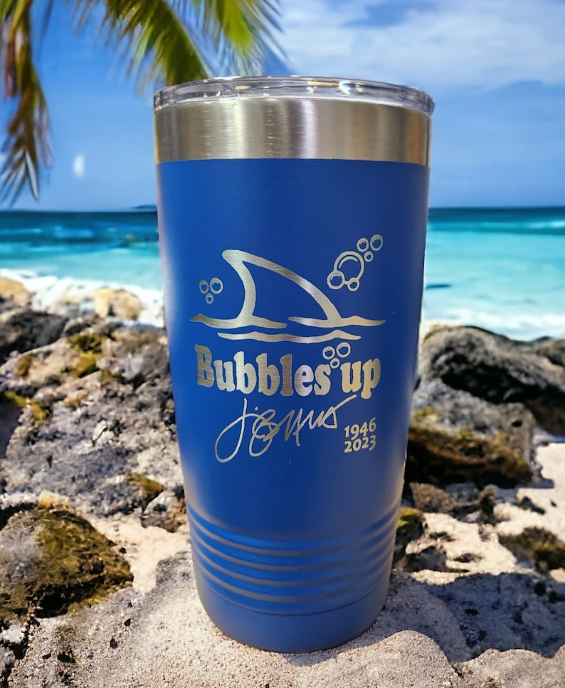 May include: A blue stainless steel tumbler with a shark fin and bubbles design. The tumbler is engraved with the text "Bubbles Up" and a signature with the dates 1946 and 2023.