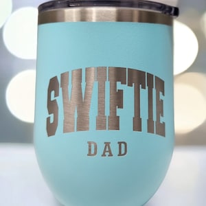 May include: A light blue stainless steel tumbler with a silver inscription that reads "Swiftie Dad".