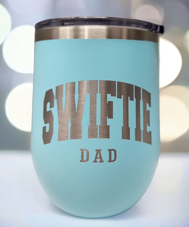 May include: A light blue stainless steel tumbler with a silver inscription that reads "Swiftie Dad".