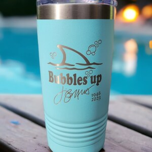 May include: A light blue stainless steel tumbler with a shark fin and bubbles graphic. The text "Bubbles Up" is printed on the tumbler, along with a signature and the dates "1946 2023".