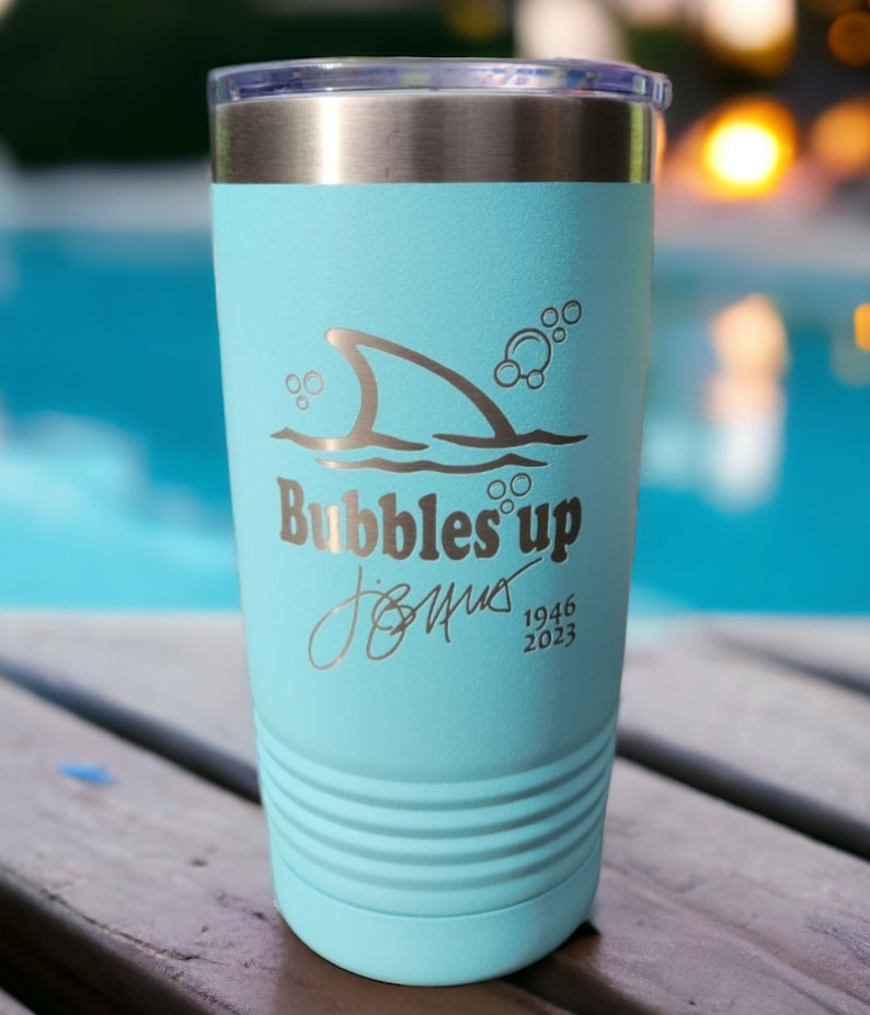 May include: A light blue stainless steel tumbler with a shark fin and bubbles graphic. The text "Bubbles Up" is printed on the tumbler, along with a signature and the dates "1946 2023".