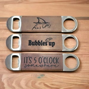 May include: Three brown leather bottle openers with metal tops. The top bottle opener has a shark fin design and the text "Fins Up!" The middle bottle opener has the text "Bubbles Up" and the bottom bottle opener has the text "It's 5 o'clock somewhere".