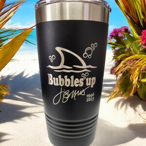 May include: Black stainless steel tumbler with a shark fin and bubbles graphic. The text "Bubbles' up" and a signature with the dates 1946 and 2023 are engraved on the tumbler.