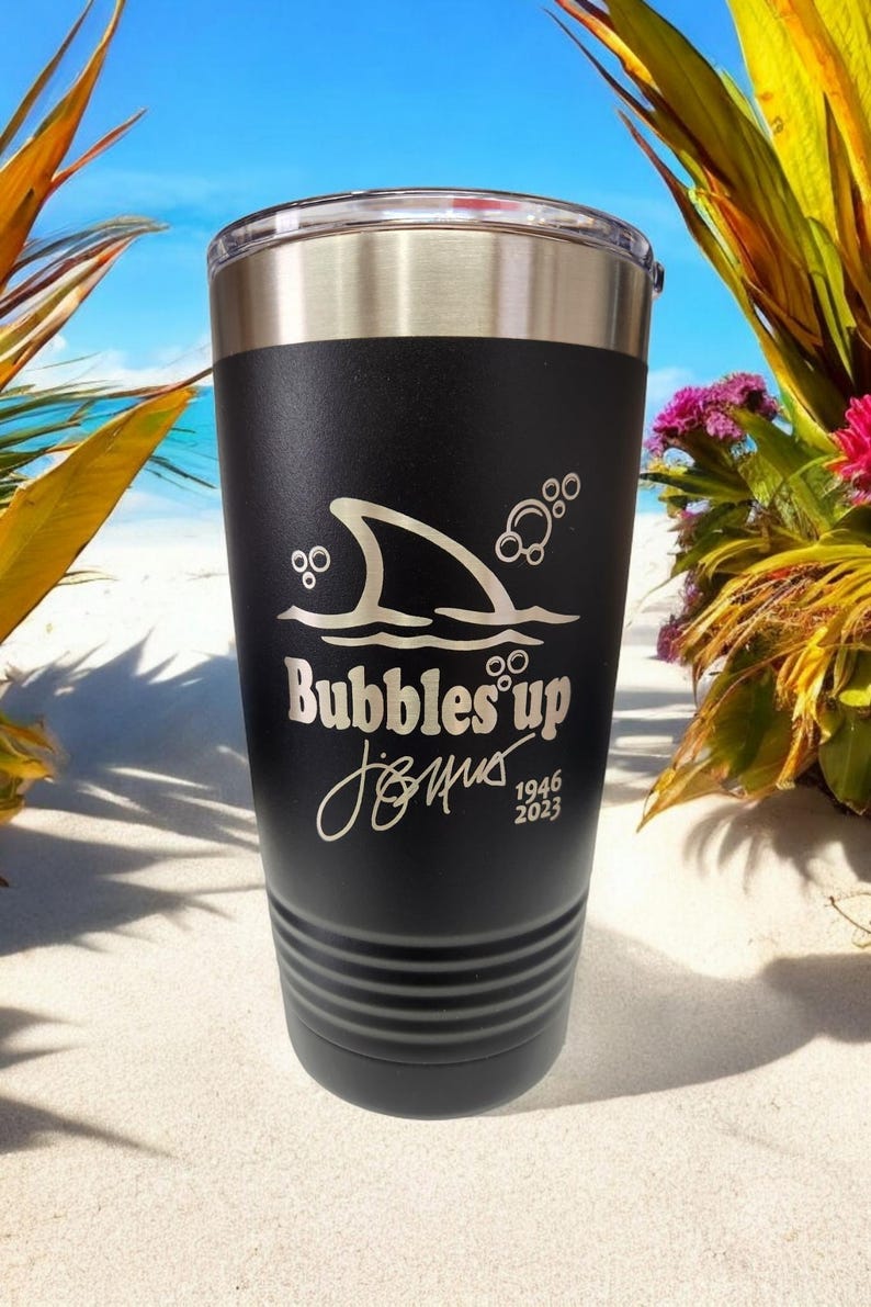 May include: Black stainless steel tumbler with a shark fin and bubbles graphic. The text "Bubbles' up" and a signature with the dates 1946 and 2023 are engraved on the tumbler.