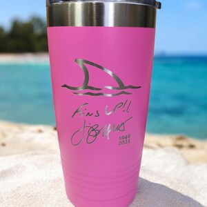 May include: A pink insulated tumbler with a silver lid. The tumbler features a shark fin graphic and the text "Fins Up!!" with a signature and the dates 1946 and 2023. The background shows a beach and ocean.